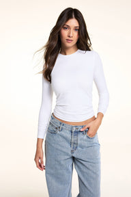 In a bright studio setting, Laura wears the Bliss Long-Sleeve Tee in White with light-wash jeans, standing with one hand in her pocket.