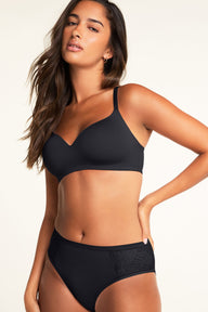 In a warm white studio setting, Kushi wears the Base Wirefree T-Shirt Bra in Black, showcasing its smooth cups, V-neckline, and slim adjustable straps, paired with the Bliss Lace Highwaist Brief in Black with decorative lace side panels.