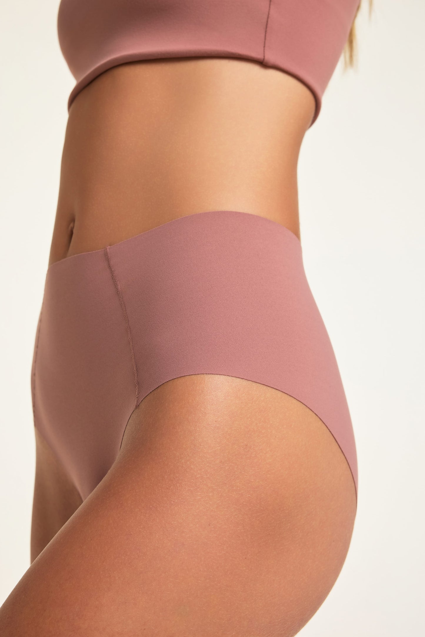 In a studio setting, Holly wears the No-Show Highwaist Brief in Plum, shown from the side to highlight the high-waisted cut and smooth, raw-edge finish.