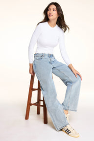 In a bright studio setting, Laura wears the Bliss Long-Sleeve Tee in White with light-wash wide-leg jeans, seated on a wooden stool and looking to the side.