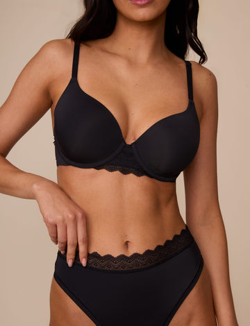 On a neutral studio backdrop, Marcela wears the Base Lace T-Shirt Bra in Black with matching underwear. The close-up front view highlights the smooth molded cups, underwire support, adjustable straps, and lace detail at the center gore and waistband. sizing: Marcela wears size 32D
