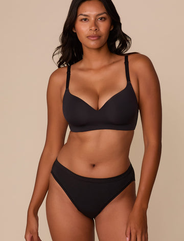 On a neutral beige backdrop, Victoria wears the Base Wirefree T-Shirt Bra in Black with matching underwear, showcasing the smooth molded cups, wireless design, and adjustable straps. sizing: Victoria wears size 32DD