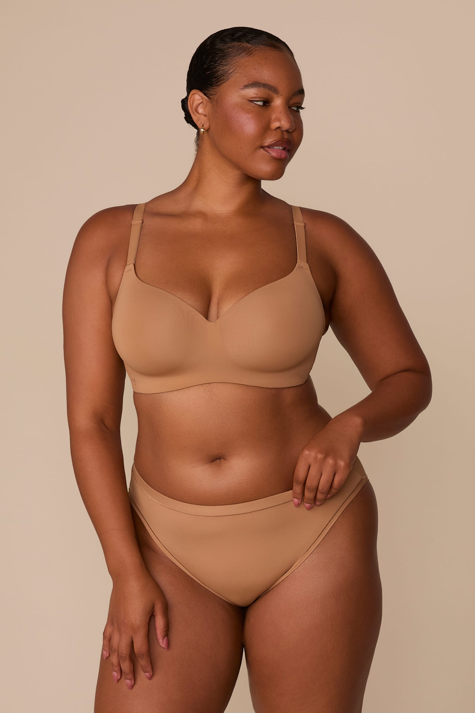 On a neutral beige backdrop, Zarria wears the Base Wirefree T-Shirt Bra in Tan with matching underwear, showcasing the smooth molded cups, wireless design, and adjustable straps.  sizing: Zarria wears size 38D