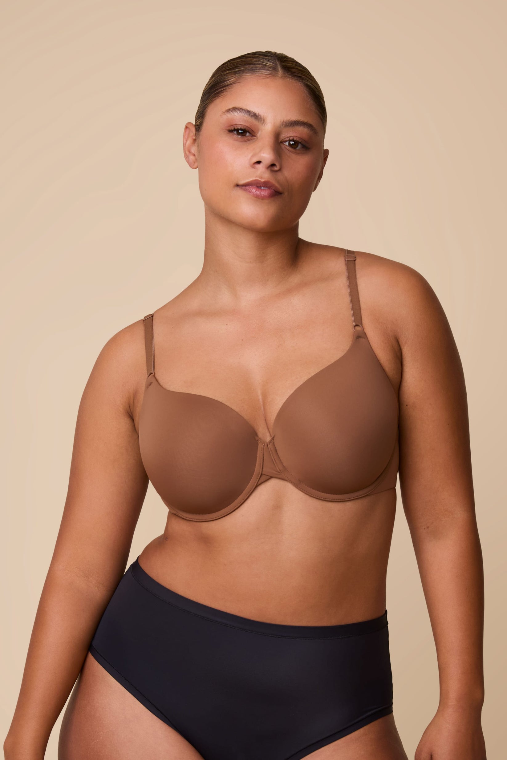 On a neutral beige backdrop, India wears the Base T-Shirt Bra in Brown, showing the lightly lined underwire cups, smooth full-coverage silhouette, and front-adjusting straps.  sizing: India wears size Large