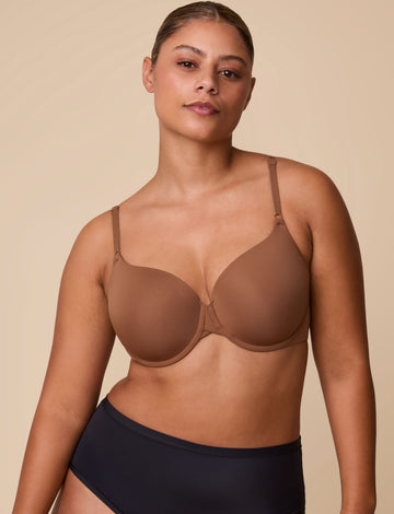 On a neutral beige backdrop, India wears the Base T-Shirt Bra in Brown, showing the lightly lined underwire cups, smooth full-coverage silhouette, and front-adjusting straps.  sizing: India wears size Large