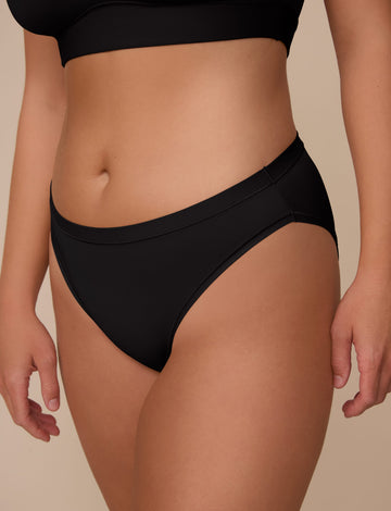 On a neutral beige backdrop, Sonnie wears the Bliss Bikini in Black, shown in a close-up front view highlighting the low-rise silhouette, smooth waistband, and soft fabric.  sizing: Sonnie wears size Large