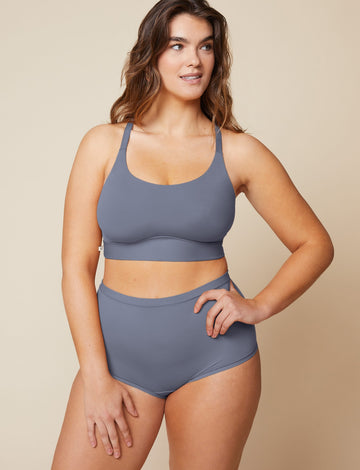 On a neutral beige backdrop, Summerlyn wears the Bliss Boyshort in Steel with a coordinating Bliss bralette, showcasing the high-rise waistband and smooth, no-dig silhouette while standing with one hand on her hip.  sizing: Summerlyn wears size Large