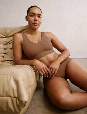 In a softly lit living room, Amber wears the Bliss Bralette in Brown, reclining against a cushioned chair. The bralette features a scoop neckline, wide straps, and a smooth, supportive underband. sizing: Amber wears size Medium