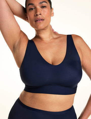 On a light studio backdrop, Jennifer wears the Bliss Bralette in Navy with matching underwear. She raises one arm overhead, showing the bralette’s V-neckline, wide wireless band, and smooth microfiber fabric. sizing: Jennifer wears size XL