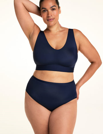 On a light studio backdrop, Jennifer wears the Bliss Highwaist in Navy with a matching navy bralette. Standing with one arm raised behind her head, she showcases the smooth high-rise waistband and full-coverage silhouette. sizing: Laura wears size Large

