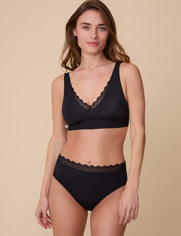 On a neutral beige backdrop, Gracie wears the Bliss Lace Bralette in Black with coordinating underwear, showcasing the deep V-neckline with scalloped lace trim, smooth fabric, and supportive underband in a full-body front view. sizing: Gracie wears size Small