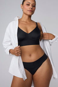 On a light gray studio backdrop, India wears the Bliss Low Rise Bikini in Black with a white button-down shirt layered over the matching Bliss Bralette in Black. The front view highlights the low-rise waistband, smooth fabric, and bikini silhouette. sizing: India wears size Large