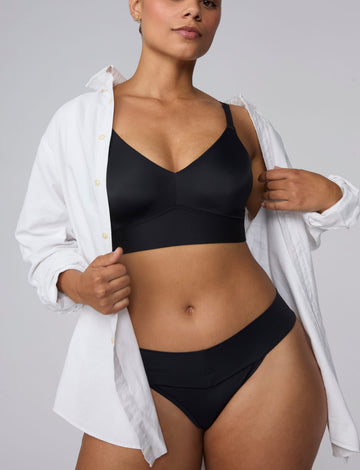 On a light gray studio backdrop, India wears the Bliss Low Rise Bikini in Black with a white button-down shirt layered over the matching Bliss Bralette in Black. The front view highlights the low-rise waistband, smooth fabric, and bikini silhouette. sizing: India wears size Large
