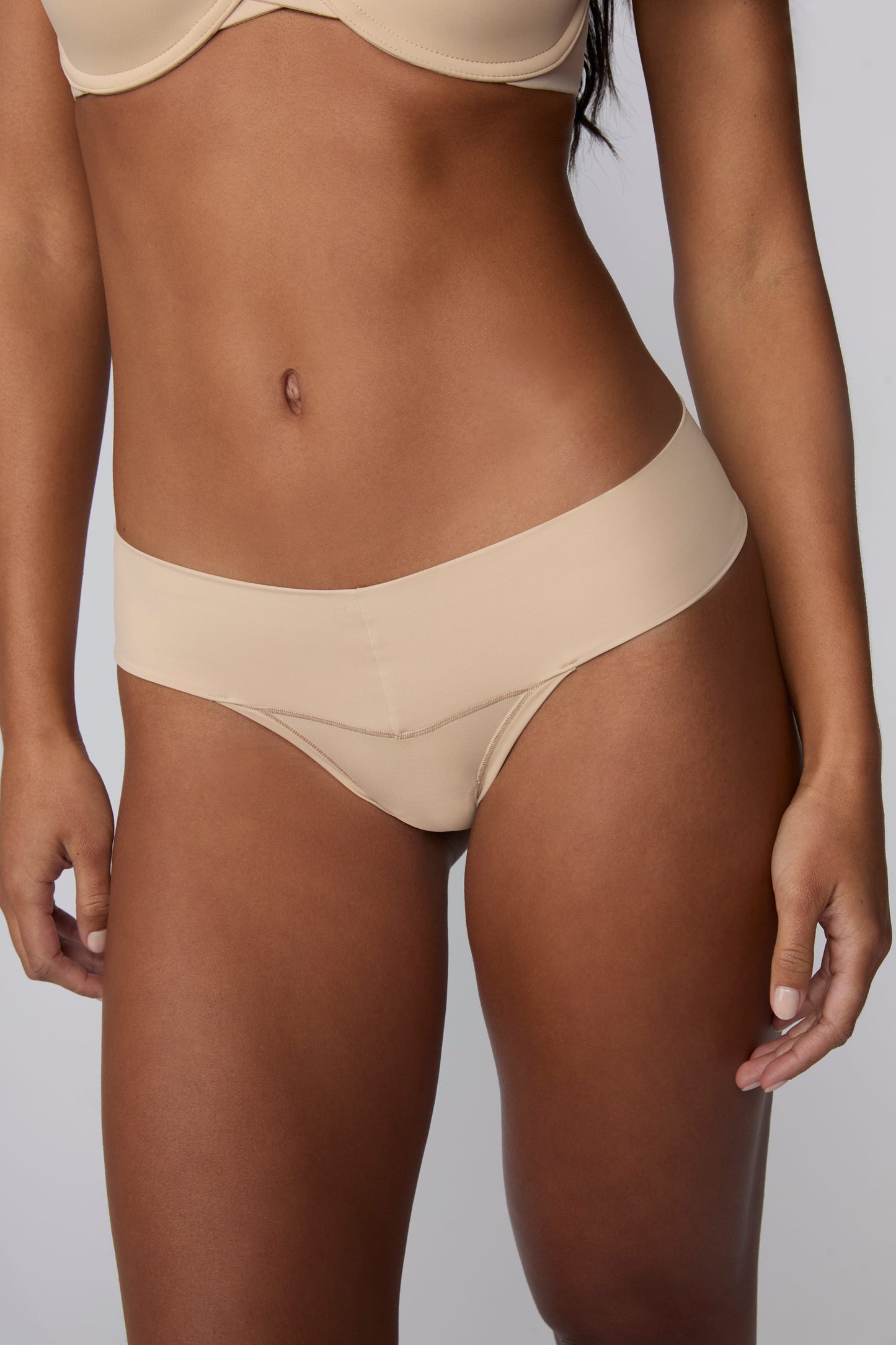 On a light gray studio backdrop, Amber wears the Bliss Low Rise Thong in Beige with the matching Base Strapless Bra in Beige. A close-up front view highlights the low-rise waistband, smooth fabric, and minimal-coverage thong silhouette.