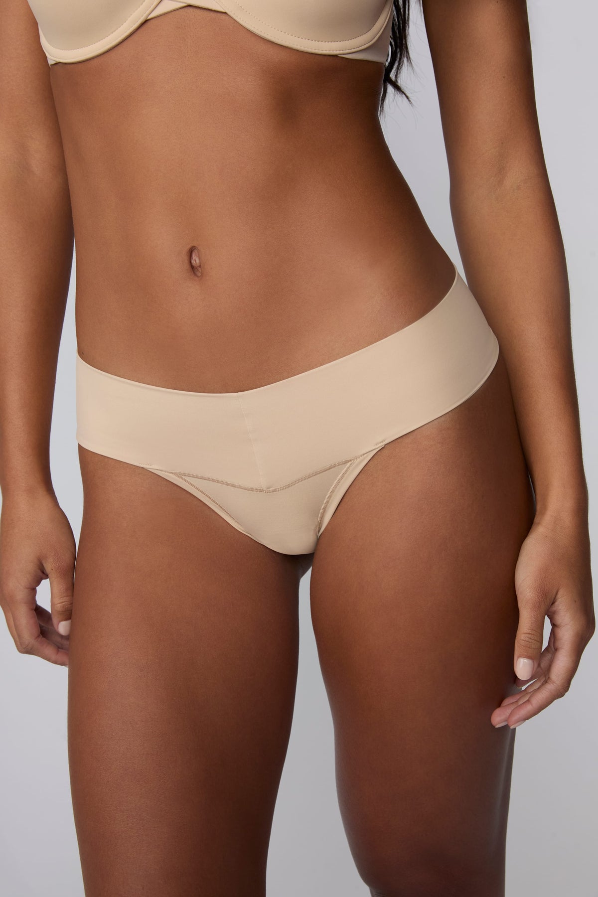 On a light gray studio backdrop, Amber wears the Bliss Low Rise Thong in Beige with the matching Base Strapless Bra in Beige. A close-up front view highlights the low-rise waistband, smooth fabric, and minimal-coverage thong silhouette.