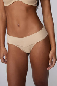 On a light gray studio backdrop, Amber wears the Bliss Low Rise Thong in Beige with the matching Base Strapless Bra in Beige. A close-up front view highlights the low-rise waistband, smooth fabric, and minimal-coverage thong silhouette.