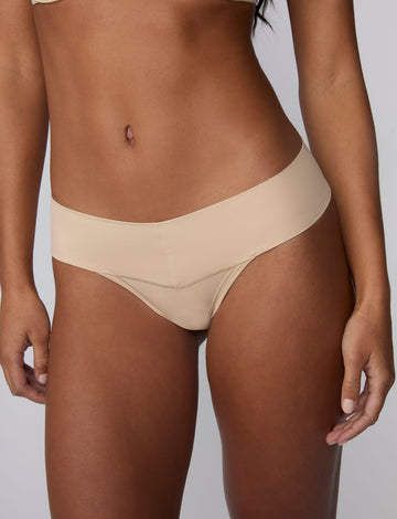 On a light gray studio backdrop, Amber wears the Bliss Low Rise Thong in Beige with the matching Base Strapless Bra in Beige. A close-up front view highlights the low-rise waistband, smooth fabric, and minimal-coverage thong silhouette.