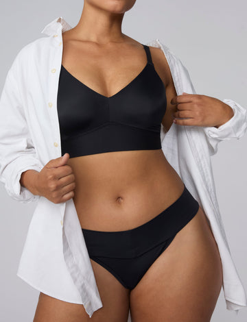 On a light gray studio backdrop, India wears the Bliss Low Rise Thong in Black with the matching Bliss Bralette in Black. An open white button-down shirt frames the look, while the front view highlights the low-rise waistband and minimal-coverage thong silhouette. sizing: India wears size Large