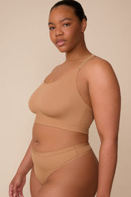 On a neutral beige backdrop, Zarria wears the Bliss Racerback Bralette in Tan with coordinating underwear, highlighting the smooth scoop neckline, adjustable straps, and supportive underband in a side-facing pose.  sizing: Zarria wears size XL