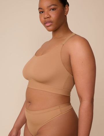 On a neutral beige backdrop, Zarria wears the Bliss Racerback Bralette in Tan with coordinating underwear, highlighting the smooth scoop neckline, adjustable straps, and supportive underband in a side-facing pose.  sizing: Zarria wears size XL