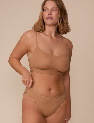 On a neutral beige backdrop, Sonnie wears the Bliss Scoop Adjustable Bralette in Tan with matching underwear, highlighting the scoop neckline, adjustable straps, and smooth underband while gently adjusting the waistband.  sizing: Sonnie wears size XL