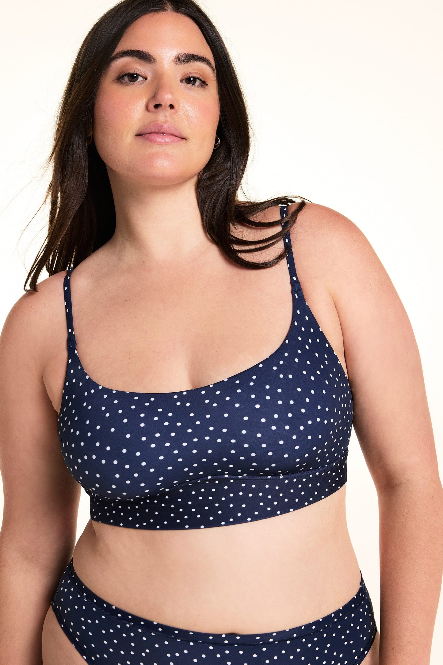On a light studio backdrop, Alessandra wears the Bliss Scoop Bralette in Navy Dot with matching Navy Dot underwear. The close-up image highlights the scoop neckline, adjustable straps, wide underband, and allover white dot print. sizing: Alessandra wears size Large