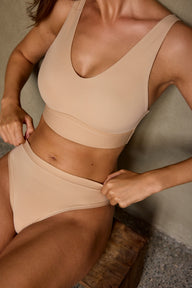 In a warm outdoor setting, Laura wears the Bliss Thong in Beige, paired with the Bliss Bralette in Beige, with hands at the thong waistband. sizing: Laura wears size Small