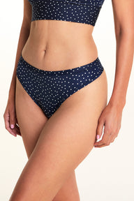 On a light studio backdrop, Meagan wears the Bliss Thong in Navy Dot. The close-up image highlights the low-rise waistband, thong silhouette, and allover white dot print. sizing: Meagan wears size Medium
