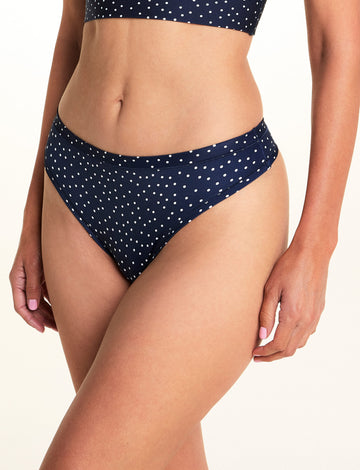 On a light studio backdrop, Meagan wears the Bliss Thong in Navy Dot. The close-up image highlights the low-rise waistband, thong silhouette, and allover white dot print. sizing: Meagan wears size Medium
