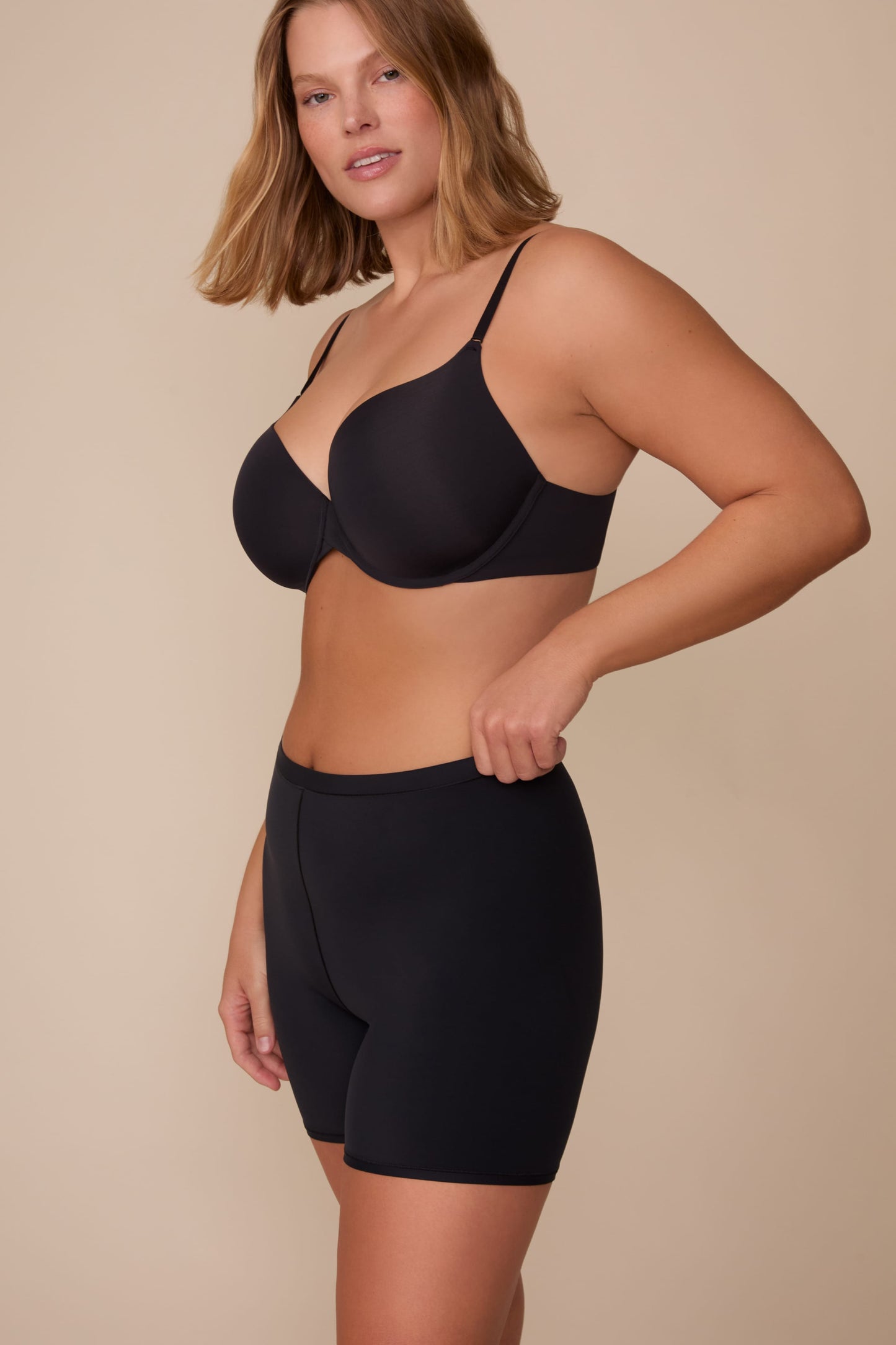 On a neutral studio backdrop, Sonnie wears the Bliss Under Short in Black with a matching black bra. The side-front view highlights the smooth, mid-rise waistband and thigh-length silhouette designed for comfortable coverage under clothing. sizing: Sonnie wears size Large