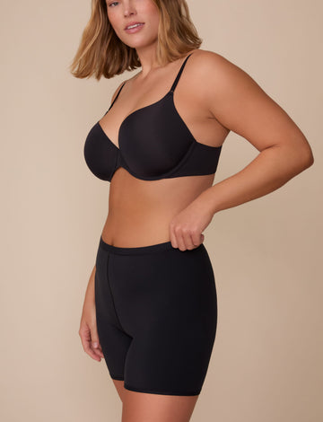 On a neutral studio backdrop, Sonnie wears the Bliss Under Short in Black with a matching black bra. The side-front view highlights the smooth, mid-rise waistband and thigh-length silhouette designed for comfortable coverage under clothing. sizing: Sonnie wears size Large