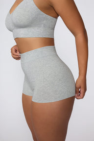 On a light gray studio backdrop, India wears the Cloud Cotton Boyshort in Heather Grey. The side view highlights the high-rise waistband, full-coverage boyshort silhouette, and soft cotton fabric. sizing: India wears size Large