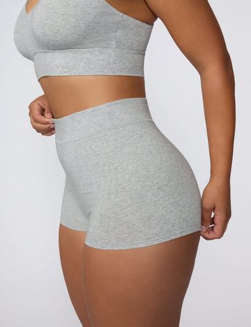 On a light gray studio backdrop, India wears the Cloud Cotton Boyshort in Heather Grey. The side view highlights the high-rise waistband, full-coverage boyshort silhouette, and soft cotton fabric. sizing: India wears size Large