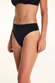 Against a white studio backdrop, Alexis wears the Cloud Cotton Highwaist Thong in Black, shown in a side angle to highlight the high-rise silhouette and smooth cotton fabric. sizing: Alexis wears size Small