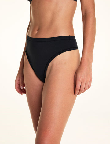 Against a white studio backdrop, Alexis wears the Cloud Cotton Highwaist Thong in Black, shown in a side angle to highlight the high-rise silhouette and smooth cotton fabric. sizing: Alexis wears size Small