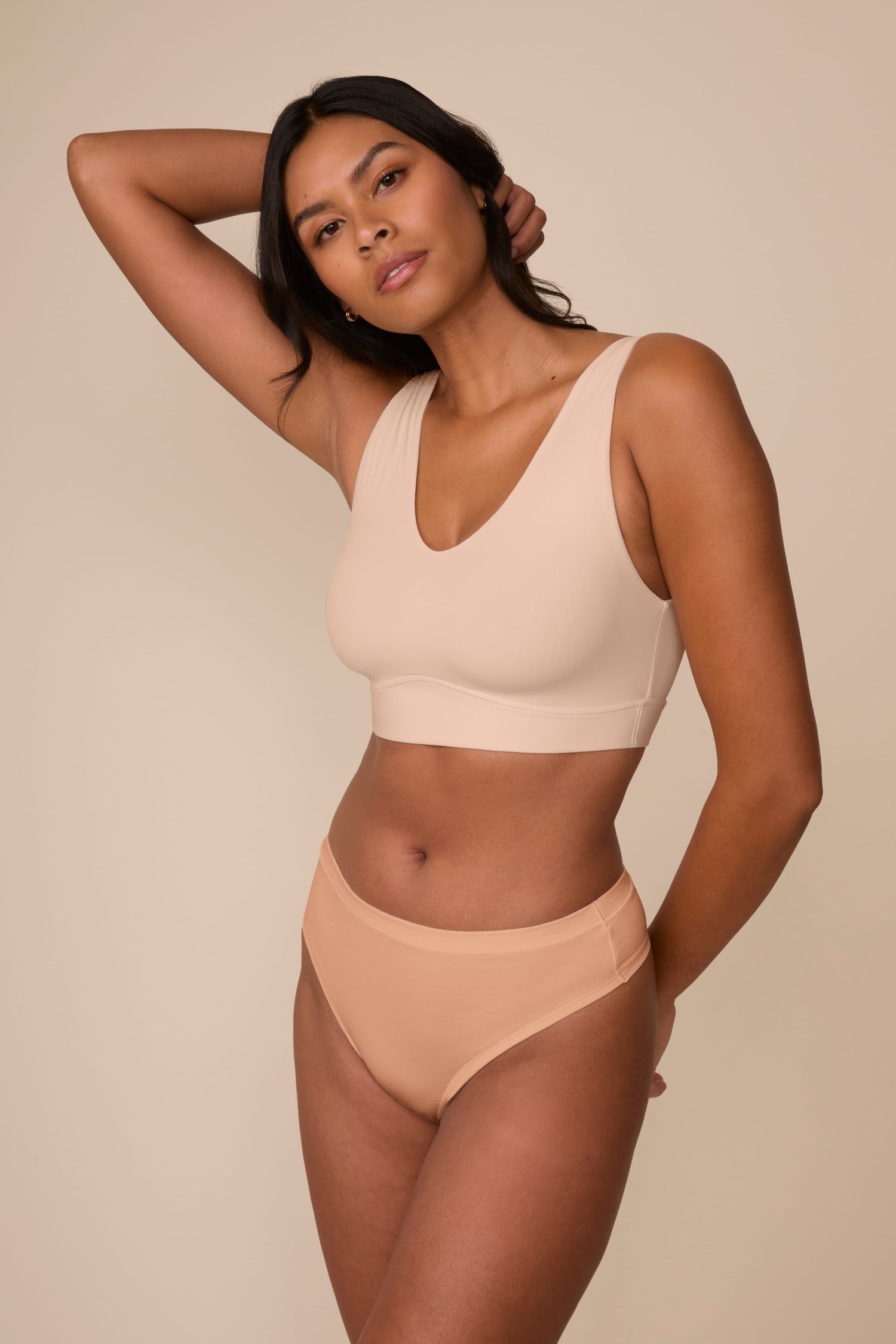 On a neutral beige backdrop, Victoria wears the Cloud Cotton Highwaist Thong in Dune with a coordinating bralette, showcasing the high-rise waistband and smooth cotton fabric while posing with one arm behind her head.  sizing: Victoria wears size Medium