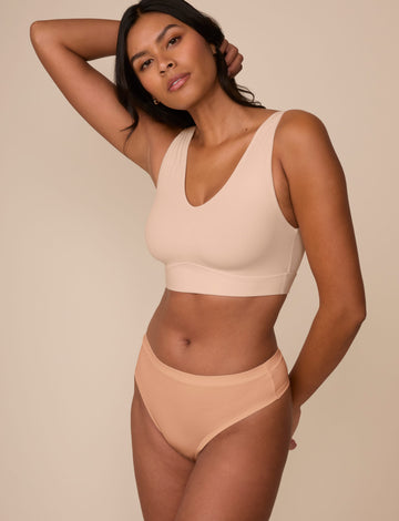 On a neutral beige backdrop, Victoria wears the Cloud Cotton Highwaist Thong in Dune with a coordinating bralette, showcasing the high-rise waistband and smooth cotton fabric while posing with one arm behind her head.  sizing: Victoria wears size Medium