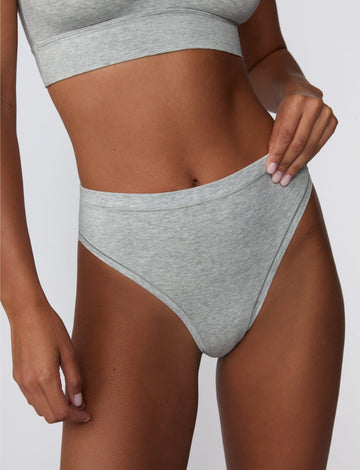 On a light gray studio backdrop, Courtney wears the Cloud Cotton Highwaist Thong in Heather Grey. The close-up front view highlights the high-rise waistband, soft cotton fabric, and high-leg thong silhouette. sizing: Courtney wears size Small