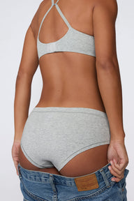 On a light gray studio backdrop, Amber wears the Cloud Cotton Hiphugger in Heather Grey with the matching bra. The back view highlights the hiphugger silhouette, low-rise waistband, full back coverage, and soft cotton fabric. A pair of jeans is lowered around the hips. sizing: Amber wears size XS