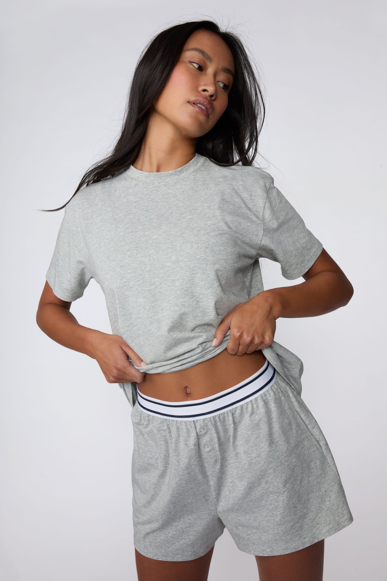On a light gray studio backdrop, Amber wears the Cloud Cotton Pajama Shirt in Heather Grey with matching pajama shorts. She lifts the hem slightly to reveal the striped elastic waistband, highlighting the relaxed crew neckline, short sleeves, and soft cotton fabric. sizing: Amber wears size XS