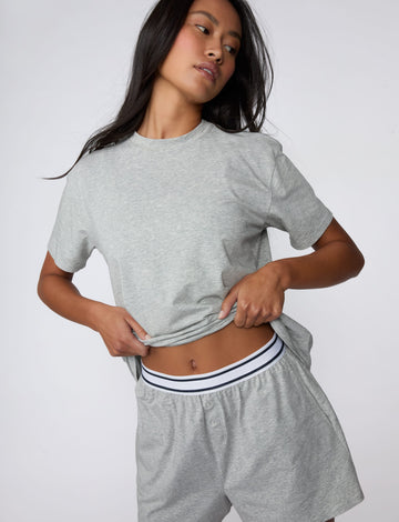 On a light gray studio backdrop, Amber wears the Cloud Cotton Pajama Shirt in Heather Grey with matching pajama shorts. She lifts the hem slightly to reveal the striped elastic waistband, highlighting the relaxed crew neckline, short sleeves, and soft cotton fabric. sizing: Amber wears size XS