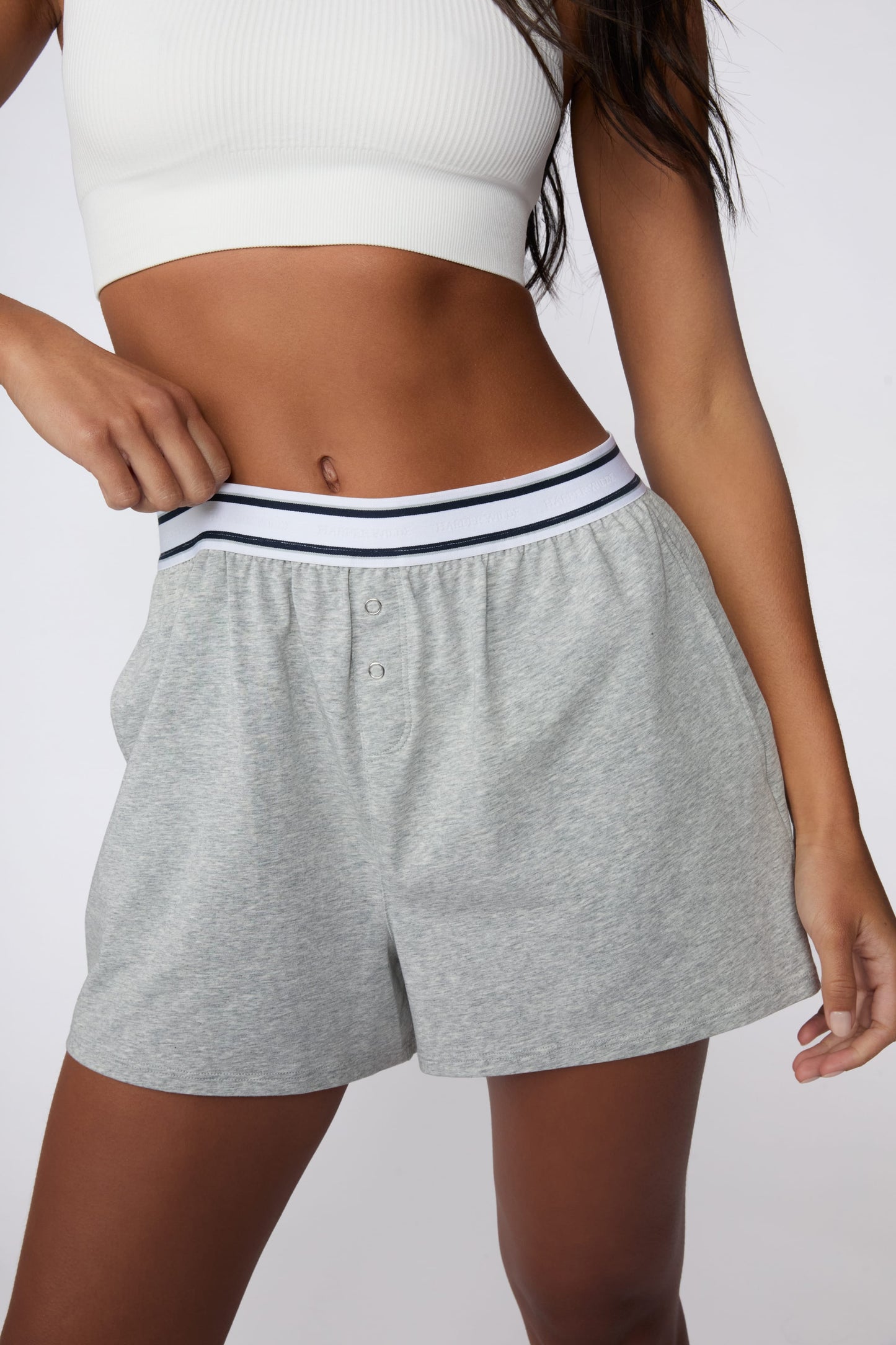 On a light gray studio backdrop, Amber wears the Cloud Cotton Pajama Short in Heather Grey with a white ribbed bralette. She gently pulls the striped elastic waistband to highlight the relaxed fit, soft cotton fabric, and functional snap-front detail.  sizing: Amber wears size XS