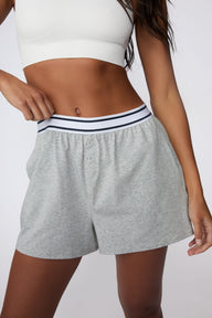 On a light gray studio backdrop, Amber wears the Cloud Cotton Pajama Short in Heather Grey with a white ribbed bralette. She gently pulls the striped elastic waistband to highlight the relaxed fit, soft cotton fabric, and functional snap-front detail.  sizing: Amber wears size XS