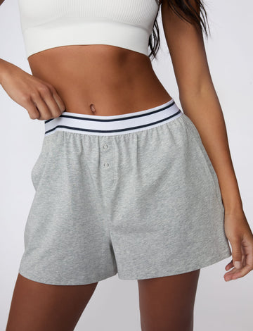 On a light gray studio backdrop, Amber wears the Cloud Cotton Pajama Short in Heather Grey with a white ribbed bralette. She gently pulls the striped elastic waistband to highlight the relaxed fit, soft cotton fabric, and functional snap-front detail.  sizing: Amber wears size XS
