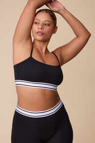 On a neutral beige backdrop, India wears the Cloud Cotton Retro Bandeau Bralette in Black with matching shorts, highlighting the straight neckline, slim adjustable straps, and striped elastic underband. sizing: India wears size Large
