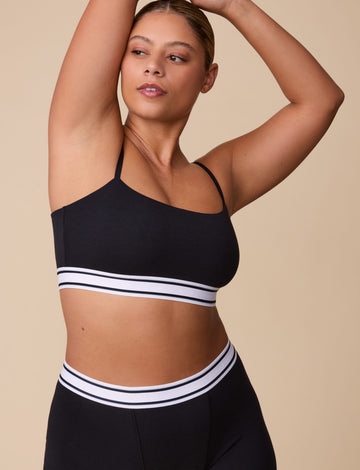 On a neutral beige backdrop, India wears the Cloud Cotton Retro Bandeau Bralette in Black with matching shorts, highlighting the straight neckline, slim adjustable straps, and striped elastic underband. sizing: India wears size Large