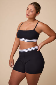 On a neutral studio backdrop, India wears the Cloud Cotton Retro Bandeau Bralette and Cloud Cotton Retro Boxer Brief in Black. She stands angled to the side, highlighting the square-neck bandeau silhouette, thin adjustable straps, and matching retro-inspired logo waistband on both pieces. The soft cotton modal fabric and full-coverage boxer brief design are visible. sizing: India wears size Small