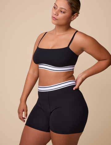 On a neutral studio backdrop, India wears the Cloud Cotton Retro Bandeau Bralette and Cloud Cotton Retro Boxer Brief in Black. She stands angled to the side, highlighting the square-neck bandeau silhouette, thin adjustable straps, and matching retro-inspired logo waistband on both pieces. The soft cotton modal fabric and full-coverage boxer brief design are visible. sizing: India wears size Small
