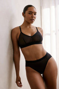 In a softly lit room, a model wears the ComfortMesh™ Highwaist Brief in Black with the matching ComfortMesh™ Triangle Bralette. The sheer mesh fabric, wide waistband, and high-waist silhouette are shown.