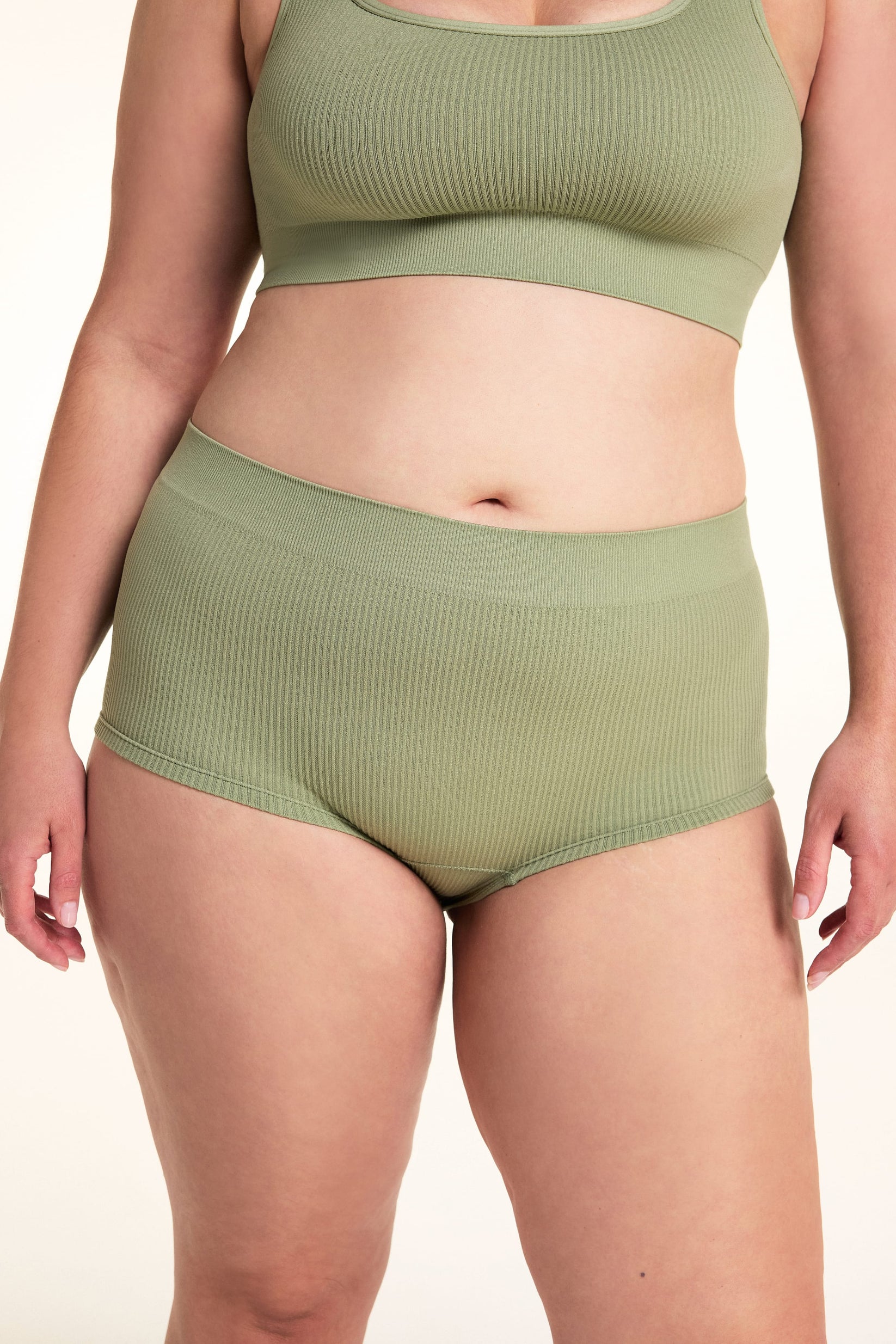 On a white studio backdrop, Alessandra wears the Lounge Rib Sleepshort in Dill with the matching Lounge Rib bralette, showing the ribbed fabric, comfortable waistband, and short silhouette.

